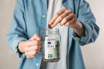 Woman putting money into tips jar on light background, closeup