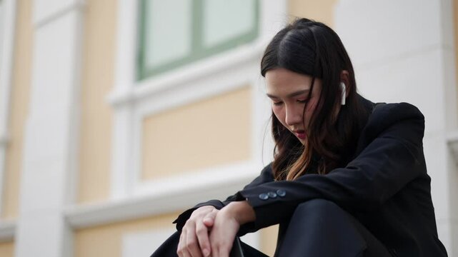 frustrated young asian woman in black suit sitting outdoors on sidewalk near office building, showing emotion of failure, sadness, and disappointment after a setback or mistake