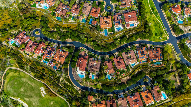 High-resolution aerial photograph showing a planned neighborhood with curved streets and luxury homes with red tile roofs and backyard swimming pools in Newport Beach, California. The community is sur