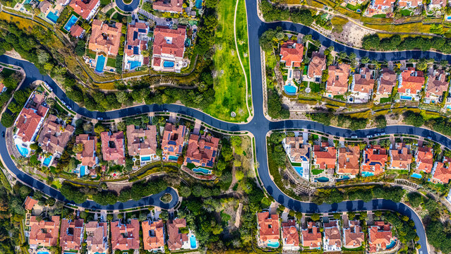 High-resolution aerial photograph showing a planned neighborhood with curved streets and luxury homes with red tile roofs and backyard swimming pools in Newport Beach, California. The community is sur