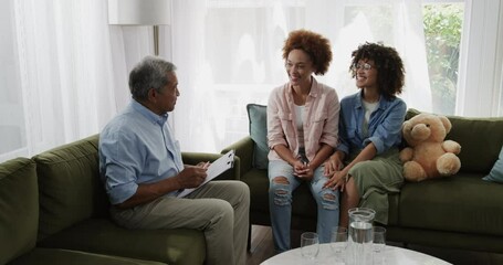 Elderly man sharing tablet with smiling same-sex diverse lesbian couple on living room sofa, at home - Powered by Adobe