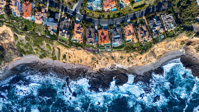 Directly overhead view of upscale homes perched along the rocky shoreline in Rancho Palos Verdes, California. The drone captures the powerful waves of the Pacific Ocean hitting the coast.