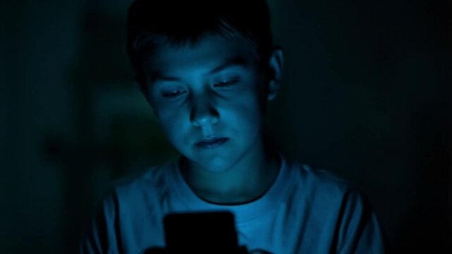 A boy is looking at a mobile phone in a dark room. Smooth repeating motion for background video animation.