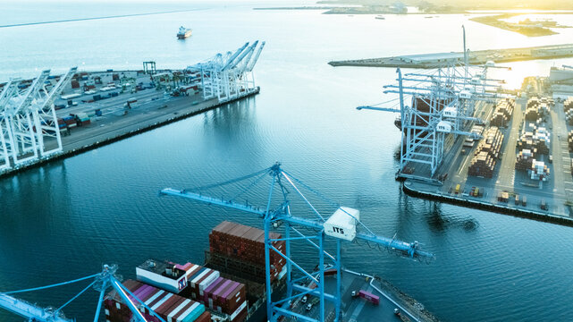 Aerial view of an expansive container terminal at sunrise, with port cranes extending over the water and cargo ships docked at berths. The morning light casts a soft glow over global trade infrastruct