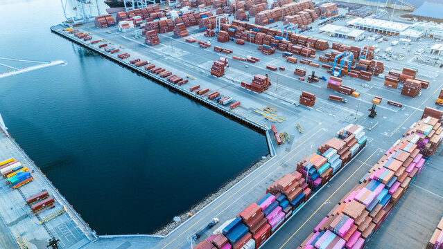 High-angle aerial view of a sprawling port complex with hundreds of shipping containers organized in rows, adjacent to the waterfront. The image captures the magnitude and complexity of modern freight