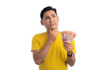 Pensive young Asian man touching his chin, holding money, looking up with doubtful expression...