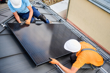 Workers building photovoltaic solar panel system on rooftop of house. Men technicians in helmets...