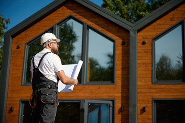 Architect hold plan. Bearded architect wearing hardhat holding project blueprints paper plans standing near comlex of new modern houses looking to something at construction site.
