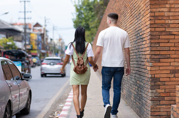 Couple of tourists walking and holding hands on a roadside sidewalk in a tourist attraction area in a city in Thailand. Fellow tourists are walking in a crowded community.