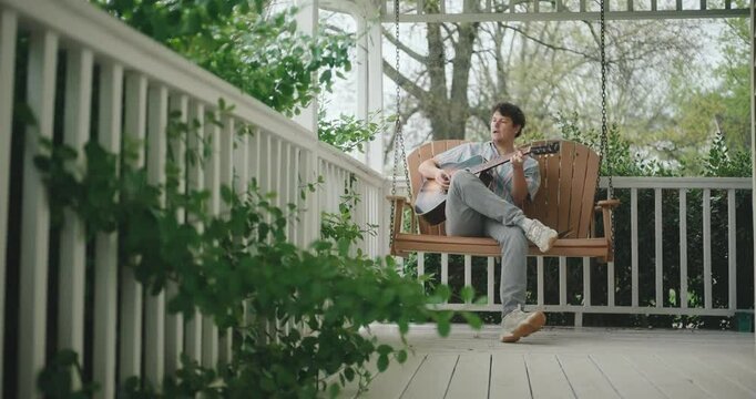 Young male musician sitting on a porch, playing guitar and writing a song