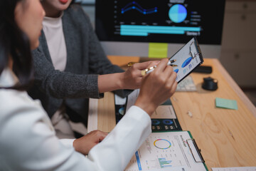 Two businesswomen are collaborating on a project, analyzing financial reports, charts, and graphs on a clipboard and computer screen, developing growth strategies for their company