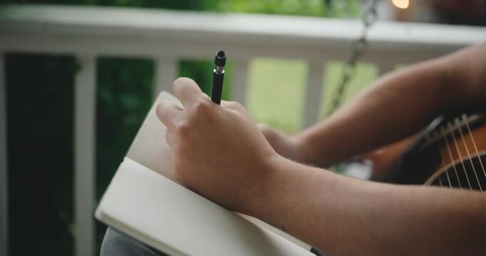 Young male musician sitting on a porch, playing guitar and writing a song