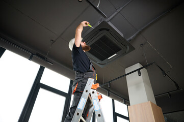 Bearded man staying on ladder installing air conditioner at the office. Ceiling mounted cassette air conditioner in modern building. Loft style. Industrial interior design. Technician servicing