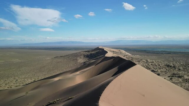 Sand dune ridge captured from above on a clear day. The scene includes a winding river behind the dune, creating a contrast between the arid landscape and the water.