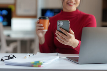 Smiling businesswoman is using a smartphone and drinking coffee while working at her desk in a modern office, with laptop and documents in the foreground