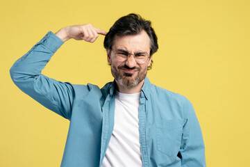 Frustrated man pointing finger at temple showing are-you-crazy gesture on yellow background