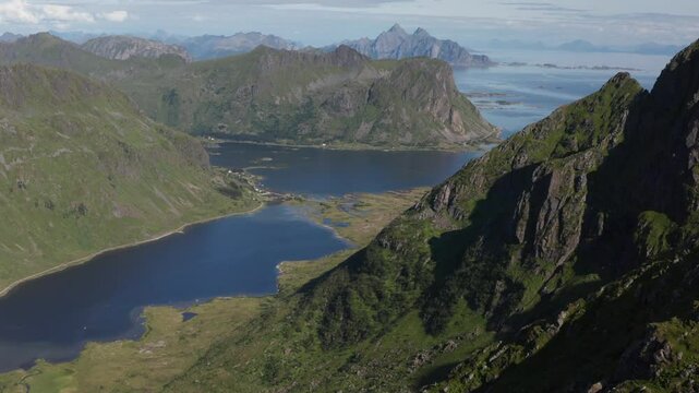 Aerial view from the summit of Justadtinden in Lofoten, Norway. Going over cliff-edge revealing dramatic mountain peaks, green hills and vast sea. Drone pushing in camera tilting down.