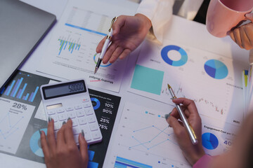 Businesswomen discussing financial data while using a calculator and pointing at various charts and graphs during an engaging collaborative meeting in a modern office setting