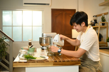 Smiling asian man using handheld electric mixer to whip cream in a stainless steel bowl