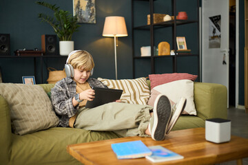 Young boy sitting on couch with headphones and tablet, focused on screen, surrounded by home decor...