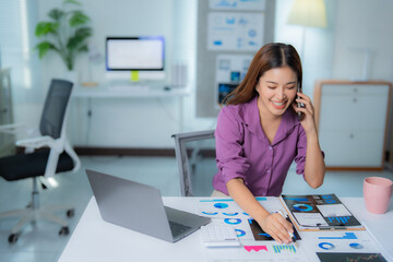 Young smiling businesswoman wearing purple shirt is having phone call and analyzing financial charts, sitting at desk in office, with laptop and calculator