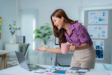 Young beautiful asian businesswoman having a video call on her laptop in a modern office while holding a cup of coffee and gesturing with her hand