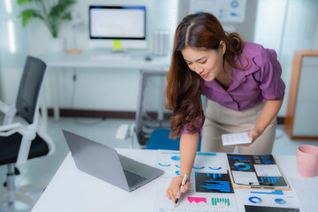 Young asian businesswoman holding calculator in hand and pointing at charts while analyzing marketing data in modern office with laptop and desktop computer