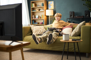 Young child with light hair sitting on spacious green sofa, engrossed in captivating movie, holding...