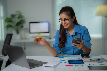 Young businesswoman holding smartphone and pointing at laptop with pen while analyzing financial...