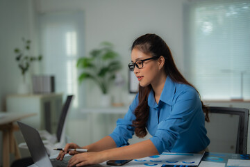 Obraz premium Young Asian businesswoman wearing glasses and blue shirt working with laptop and analyzing financial charts at office desk, holding pen in her right hand
