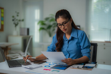 Young businesswoman wearing glasses and a blue shirt, analyzing financial data and charts while...