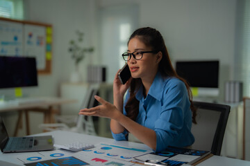Fototapeta premium Young professional woman wearing glasses and blue shirt is sitting at desk in office, talking on phone and discussing charts, using laptop and calculator