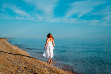 photo of dawn on the sea of ​​Azov, blue sky, blue sea, rays of dawn sun, soft sand, waves and red-haired girl standing by the shore with her back to the shore without a face in a white dress