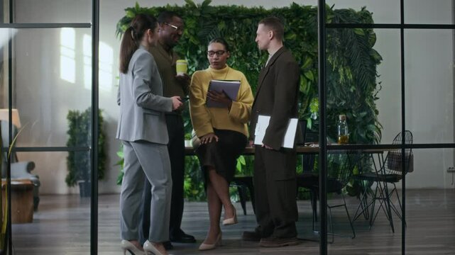 Full slowmo shot of four young diverse colleagues in smart casual clothes chatting during coffee break in modern office interior with glass partitions and lush green wall in background