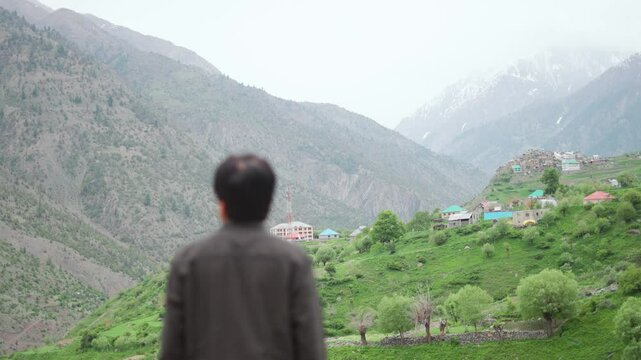 4K Rack focus shot of an Indian man looking at Triloknath village in front of the snowy Himalayan mountain peaks at Pattan valley in Lahaul, Himachal Pradesh, India. Man in front of village on cliff.