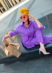 woman with craft shopping bags in the city. The concept of fashionable Consumerism and ecology. An adult Asian woman in a purple suit with red and yellow accents enjoys shopping . a sunny day.
