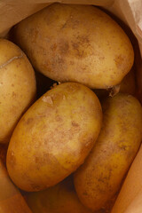 A close-up shows several unpeeled potatoes with slightly textured, light brown skin inside a brown paper bag