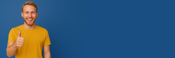 Smiling young man in a yellow t-shirt showing a thumbs up gesture against a plain blue background