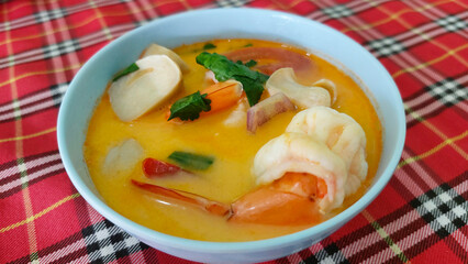 Close-up of a flavorful bowl of Tom Yum soup with fresh shrimp, mushrooms, and Thai spices, set on a red checkered tablecloth