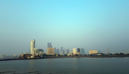 Obraz premium Early morning light casts a calm glow over the Mumbai skyline, showcasing modern buildings and tranquil water below