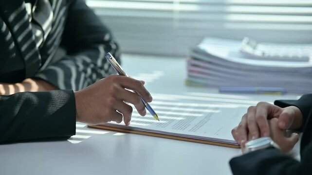 Two people are sitting at a table with a pen and a piece of paper. One of them is holding the pen and the other is looking at the paper. Scene is serious and focused