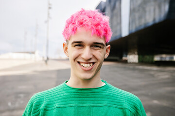 Young trendy man with pink hair laughing candidly standing outdoors. Portrait of fashionable gen z caucasian gay male at city street. People concept