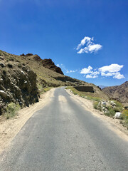 A winding road curves through the rocky terrain of Leh Ladakh, surrounded by mountains and a bright blue sky with fluffy clouds above
