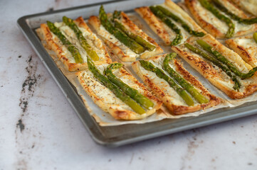 Small puff pastry tarts with green asparagus, fresh cream and parmesan. Asparagus pie with cheese on a metal tray on stone background. Spring healthy food.
