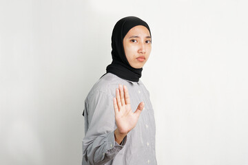 Young Asian woman raising her hand with palm facing forward in a stop gesture, expressing refusal or warning with a serious expression while standing against a plain background.