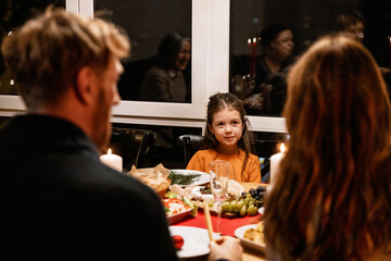 Daughter smiles at her mom, who is sitting next to dad, during Christmas dinner. Other family members at the table are reflected in the window. A concept of reunion, celebration, and family warmth.