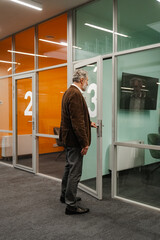 A white man stands near a glass office door marked with the number three in a modern workplace. He is waiting outside a meeting room in a professional building.