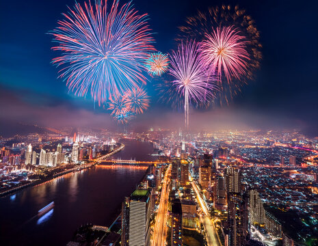 Colorful fireworks above a glowing night cityscape with river reflections, viewed from high above

