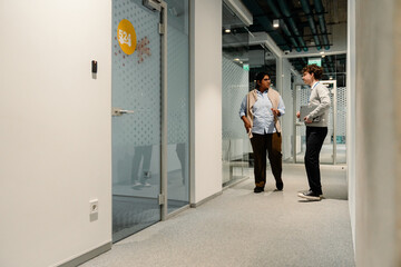 Indian woman and a White man standing and discussing in a modern indoor office hallway, holding documents and wearing business casual clothing.