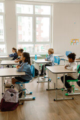 A White blonde schoolboy with his multinational group of classmates are sitting attentively in a classroom, each at their desks and wearing school uniforms in a bright, modern educational environment.